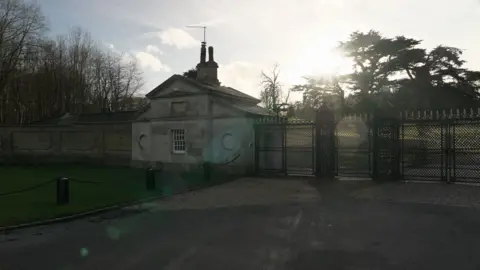 Sunlight glares across a black gate connected to a white gatehouse at the entrance of Althorp House 