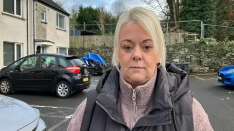 A woman with blond hair is standing beside cars and houses.