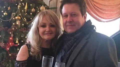 A glamourous blonde woman wearing a sequined black top, stood with her arm around a brunette man wearing a smartgrey coat and a scarf. They are both holding champaign glasses. Behind them is a Christmas tree decorated with baubles and lights.