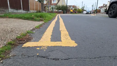 BBC Close up of double-yellow lines stretching away alongside a suburban street with cars and houses in the background
