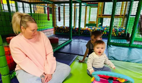Sarah Wilson A woman in a pink jumper and jeans sits in a soft play next to her two young sons. She turns to smile at them, who are both looking up at the camera. A ball pit is visible behind them.