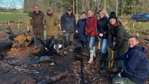 Bowood Estate A small group of smiling people, all wearing winter coats, smile at the camera besides a patch of dug-up earth. Two of the people are holding garden spades, and one person is holding what appears to be a small evergreen shrub or tree. An area of woodland can be seen in the background, as well as two trucks. 