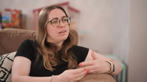 BBC Katy, who wears glasses and a black T-shirt, talks to BBC Panorama from her sofa. Toys can be seen in the background.
