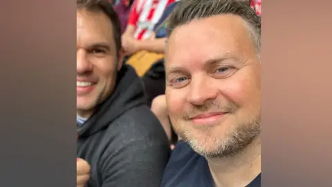 Daniel McIlwrath Daniel McIlwrath is sitting in a crowd of football fans. He is smiling in the selfie and has swept back short brown hair and is wearing a navy T-shirt.