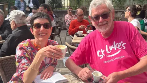 A white haired man in a bright pink t-shirt sits with a younger lady with dark short hair in an outdoor cafe.