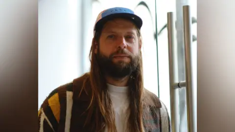 Tom Barrett A portrait of Tom who is looking at the camera with a pensive expression. He has long light brown hair and a full beard and moustache. He wears a white T-shirt, brown and mustard cardigan and a peaked cap. He is indoors.