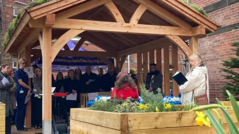A group of people gathered at a wooden summer garden house listening to a woman in a beige coat reading 