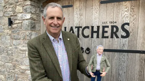 Martin is wearing a sage green tweed jacket and checked shirt, staring at the camera with a slight smile on his face. He is holding an small model of himself wearing the exact same clothing. Behind him is the word 'Thatchers' on a gate, with a wall to the side.