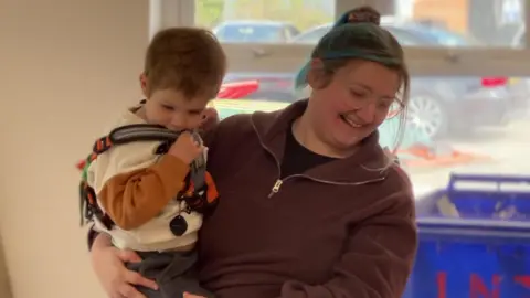 Hayleigh Woolven holds her son in her arms whilst she smiles. She has blue hair, a brown jumper and glasses, whilst her son, who is aged around 3, has a white and orange jumper on.