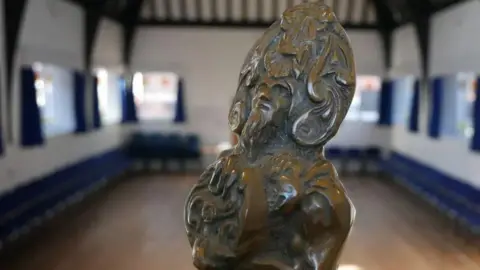 CULTURE VANNIN Close-up of the head of the RBV trophy depicting the sea god Manannan. It is in an empty hall which has wooden rafters and and a wooden floor and is lined with blue chairs positioned under its many windows.