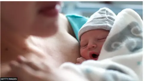 Getty Images Close-up of a mum holding her sleeping newborn baby on her chest. Baby is wearing a pale coloured hat and is wrapped in a blanket.