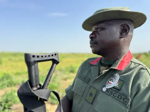 Ayo Bello / BBC Um homem com uniforme cáqui segura uma arma.