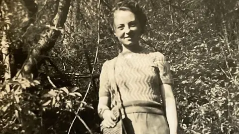 A black and white picture of Alice Moody. She is smiling at the camera.  Alice is wearing a jumper and skirt and she is holding a handbag.  Trees and bushes are in the background.