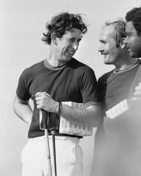 Getty Images Charles holds a polo club and chats with other players. He smiles and his hair is mussed