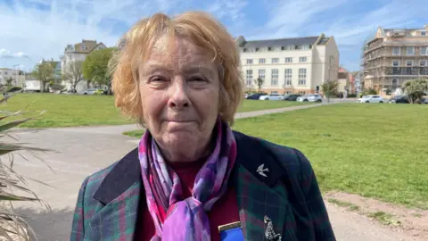 A lady wearing a purple and pink scarf with a green jacket smiles at the camera. Behind you can see a green and flats in the distance.