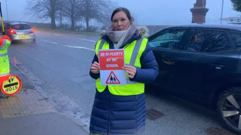 A woman wearing a long winter coat and a hi-vis jacket, stands at the side of the road and holds a road safety sign which says "20 is plenty by my school".