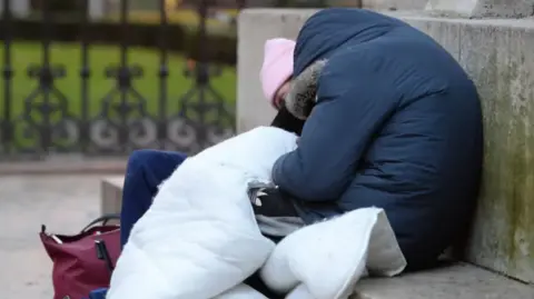 A woman rough sleeper curls up in a blue coat and a white duvet over her legs
