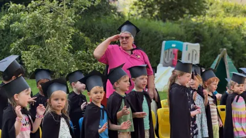 BBC Sheila Dwyer looks out behind her class of little children all wearing their graduation mortar boards and gowns