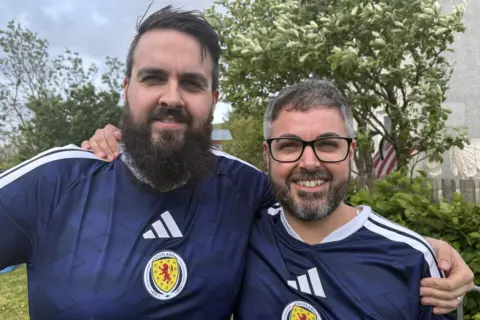 Two men with in blue Scotland shirts with their arms round each other's shoulders. Both men have brown hair and beards, and the man on the right has glasses.