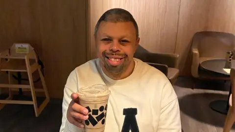 Ann-Marie Kinsman Reece Kinsman sitting in a cafe. He is wearing a white jumper, holding a coffee and is smiling. He has short hair and some facial hair.