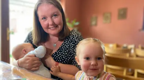 Katy, wearing a polka dot top and a chain with a light blue pendant, holds a small child chose to her chest, while another, slightly older child sits in front of them, wearing a colourful stripey top. 