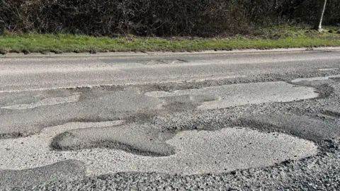 A section of large potholes scarring a road with a grass verge in the background.