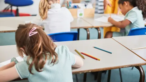 Young girls in a classroom, faces are not visible as they are photographed from behind, sat at school desks, the girl in foreground has long brown hair, pink hair clips, and is wearing a green checked top, there are pencils visible on the desk beside her 