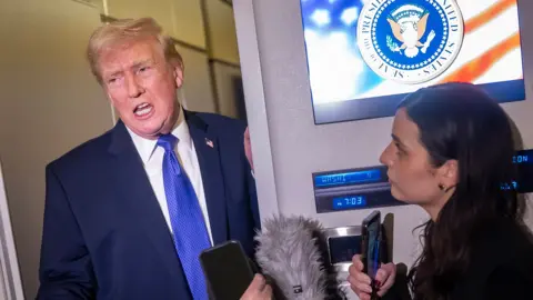 Donald Trump gestures as he speaks to reporters while wearing a suit and blue tie and holding onto a door frame next to a screen that has the president's logo on it that says 'seal of the president of the United States', on Air Force One on Monday.