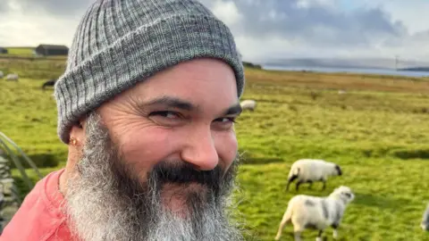 Jonathan Berner A man with long black and white beard wearing a grey knitted hat is standing in front of a green field in Shetland, with a few sheep visible behind him.