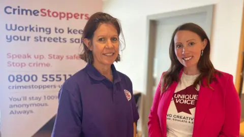 BBC Karen MacDonald (left) from Crimestoppers in purple polo shirt and Krystal Keely from Unique Voice in pink blazer standing in front of a Crimestoppers banner inside a room