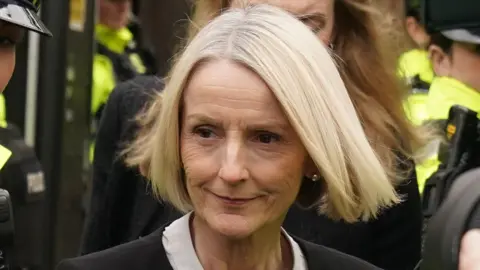 PA Media A woman in her late 50s with blonde hair wearing a black jacket and white blouse viewed from the neck up. In the background, police officers in high-vis.