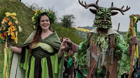 A man and a woman wearing green, with the woman holding a bunch of yellow and red flowers and the man a green mask with antlers on it, with a grassy hill behind them