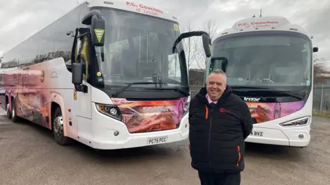A man with short grey hair, dressed in a thick black coat, smiles as he stands in front of two large coaches with big tinted windows. The bodywork is partially white and partially painted in shades of red and purple with scenes of holiday locations including mountains and ferris wheels.