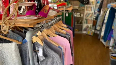 Bedford Borough Council A clothes rail in a charity or second‑hand shop displaying assorted tops on wooden hangers, with shelves of shoes and other items visible in the background.
