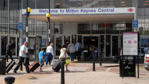 The outside of Milton Keynes rail station, with several people walking into it wheeling suitcases behind them. A sign - blue writing on white - says Welcome to Milton Keynes Central Station.