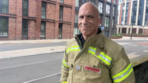 Group manager for the Avon Fire and Rescue Service, Paul Smith, smiles at the camera as he stands outside of the tower block in Old Market, Bristol City Centre. He is wearing his Avon Fire and Rescue Service uniform.