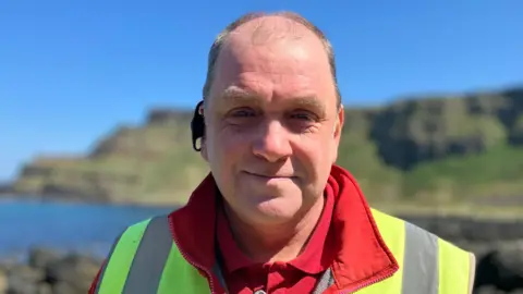 A man in a red hoodie, yellow hi vis vest with an ear piece in looks at the camera with a slight smile. He has light coloured hair.