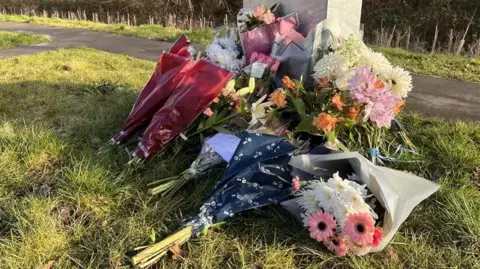 Charlie Rose/BBC Bouquets of flowers placed on a patch of grass next to a path.