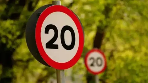 A round road sign that reads 20 in black writing on a white background with a red border. There is another in the background and a bush behind.