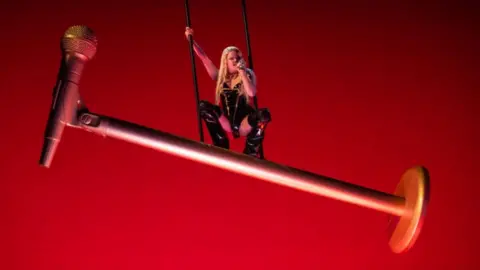 A woman sits on a giant microphone during a stage performance.