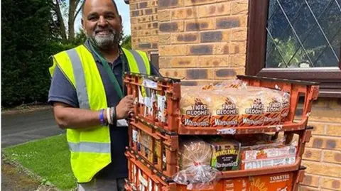 FODK A man wearing a green high-vis jacket next to crates of bread. The crates are orange and behind him is grass and a brick wall of a house. 