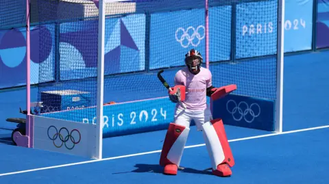 Elsa/Getty Images Miriam Pritchard of Team Great Britain reacts during the Women's Pool B match between Great Britain and Spain on day two of the Olympic Games Paris 2024 at Stade Yves Du Manoir on 28 July 2024 in Paris, France. She is in her kit and holding a hockey stick. The pitch is various shades of blue. The Olympic circles are illustrated at the side of the goal gate.