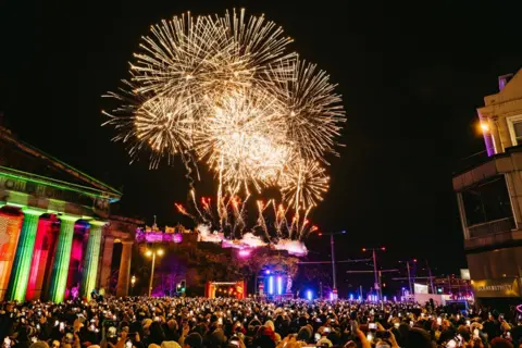 Unique Assembly Gold fireworks in a dark sky while a crowd of people below film on their phones.
