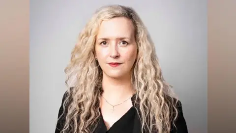Victoria Bell is wearing a black top, belt and jacket and has long blonde curly hair. She is looking straight at the camera.