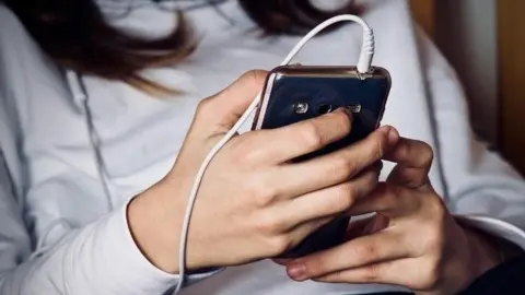 A teenage girl looks at her mobile phone, sitting and wearing a white top and black trousers