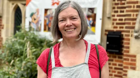 A woman wearing a pink top and dungarees smiling in front of a building