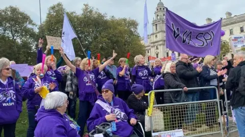 A large group of women in purple t-shirts and jumpers standing outside Parliament with banners and signs.