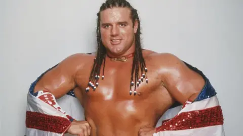 Getty Images Wrestler British Bulldog stands smiling with his hands on his hips and flexing his arms. He has long, braided hair and has a sparkly red, white and blue flag wrapped around him.