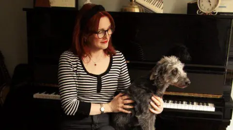 Noah Star Lou Hickey sits by her piano while holding her dog in her arms - Lou is wearing a black and white stripey top and has glasses and red hair.
