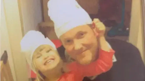 A close-up photo shows a father and daughter people indoors wearing matching white hats, standing side by side in a narrow hallway. The young girl is in a red top has an arm placed around the man’s shoulder, and both are facing the camera. The background includes a doorway and household items, with soft, slightly blurred lighting throughout the image.